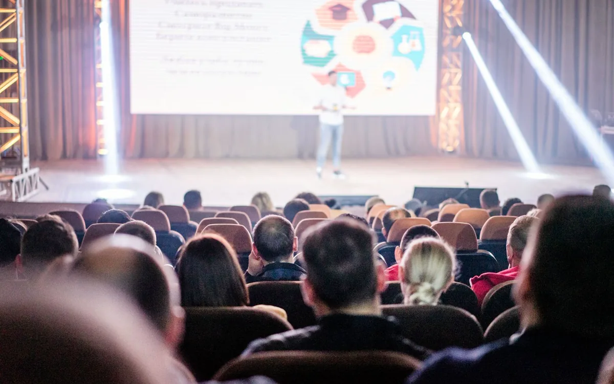 Publikum im Saal vor einer Bühne mit Leinwand und Scheinwerfern